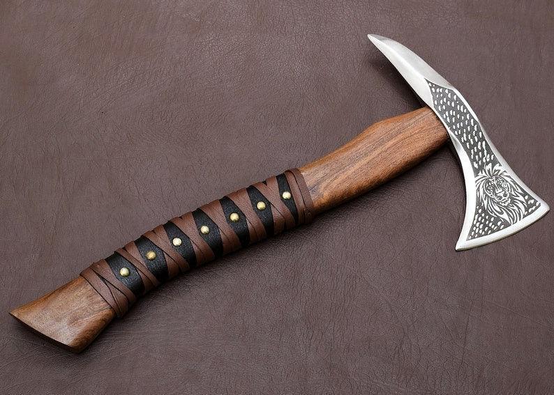 A handcrafted axe with a Corban steel blade and a Rose wood handle, featuring a decorative pattern on the blade and a leather cover.