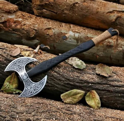 A handmade axe with a black and silver patterned head and a brown and black wrapped handle lying on a wooden surface with leaves around it.