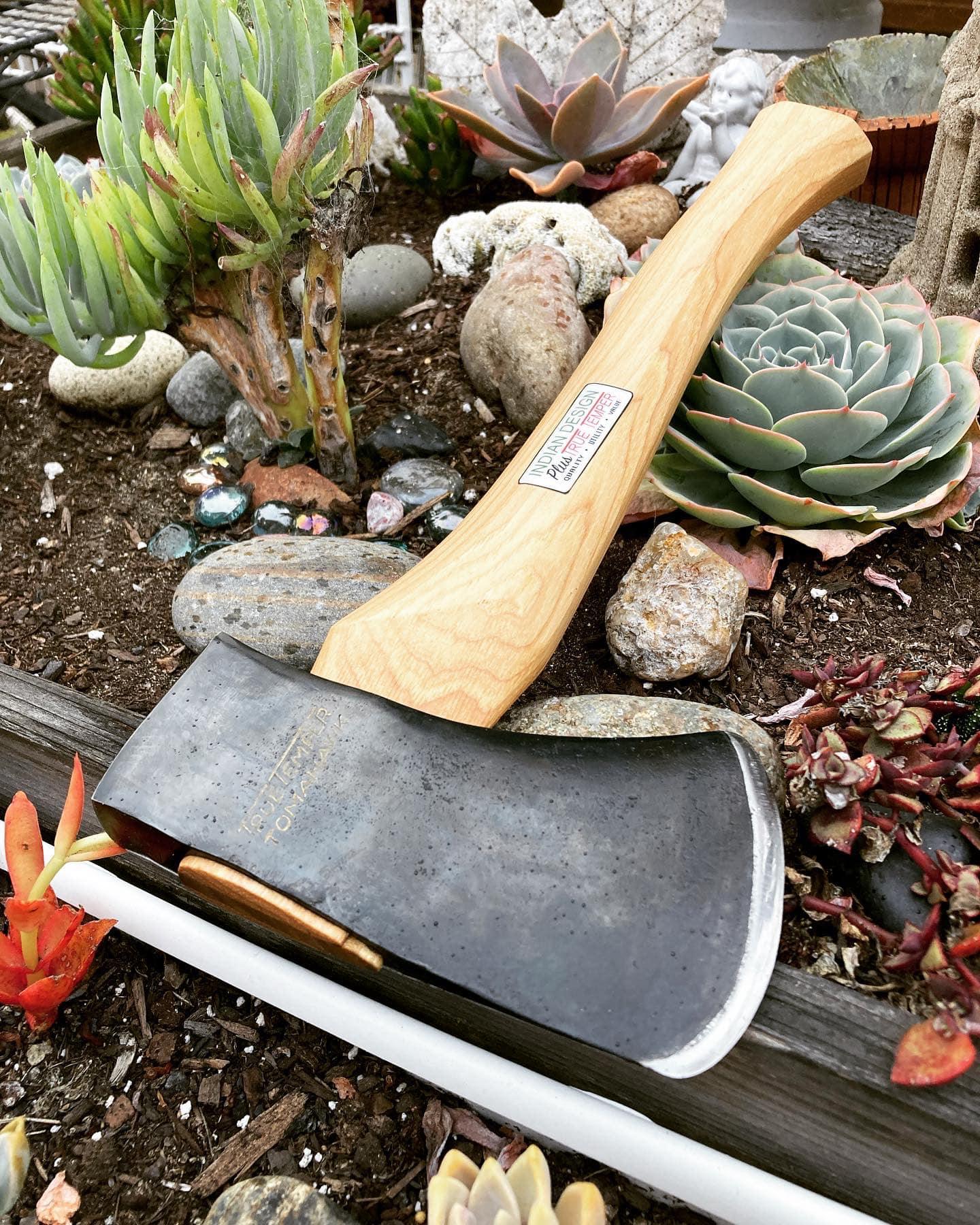 A handcrafted axe with a Corban steel head and Ash wood handle, displayed in a garden setting with a handmade leather cover placed beside it.