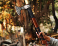 A handcrafted Viking axe with a detailed etched blade and an Ash wood handle, held by a person with a blurred natural background.