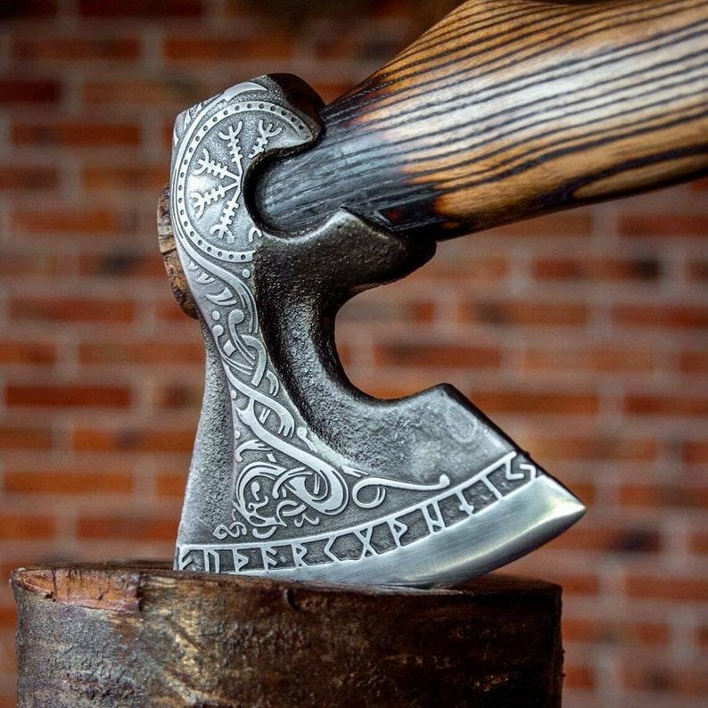 A close-up of a handcrafted axe with a detailed, engraved blade, featuring a mixture of Corban steel and wooden handle, placed against a brick wall backdrop.