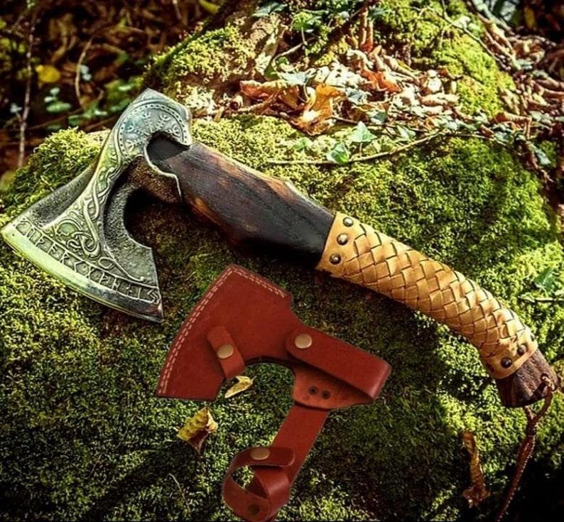 Engraved steel axe with a brown leather cover and a wooden handle, placed on a rock with a forest background.