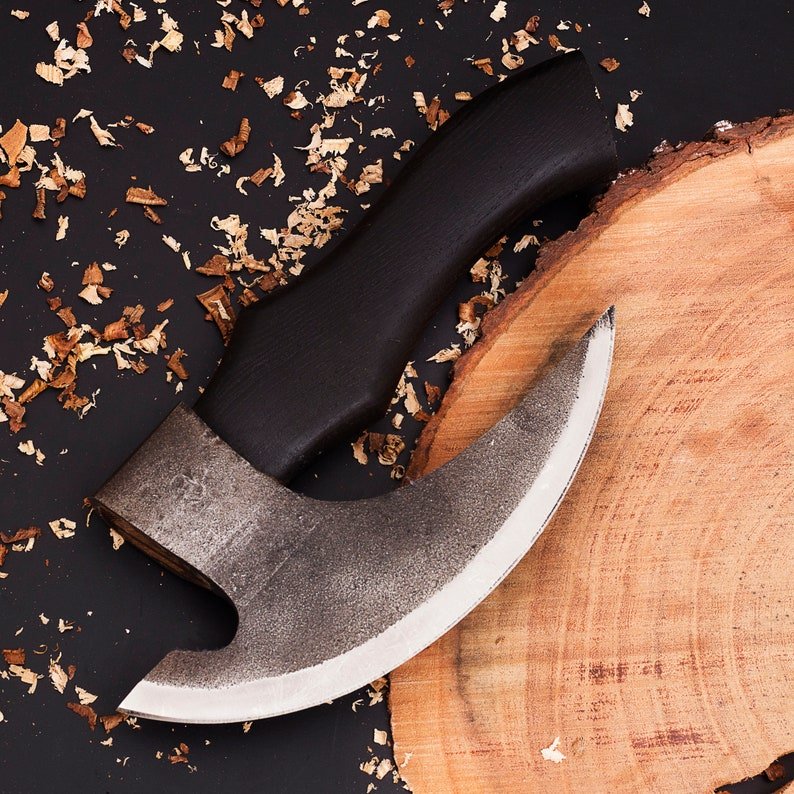 A hand forged pizza axe with a leather cover, resting on a wooden surface with sawdust scattered around, highlighting its sharp blade and wooden handle.