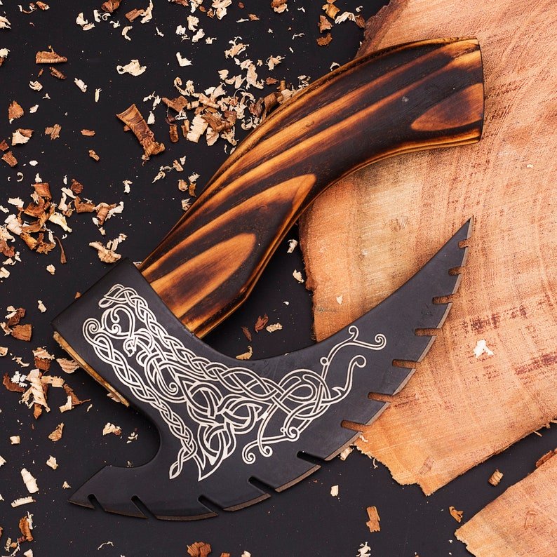 A handmade engraved pizza axe with a wooden handle and a leather cover, resting on a dark surface with wood shavings.