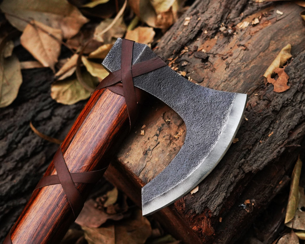 A close-up of a steel axe with a wooden handle, lying on a bed of leaves. The axe has a handmade leather cover tied around its handle.