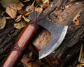 A close-up of a steel axe with a wooden handle, lying on a bed of leaves. The axe has a handmade leather cover tied around its handle.