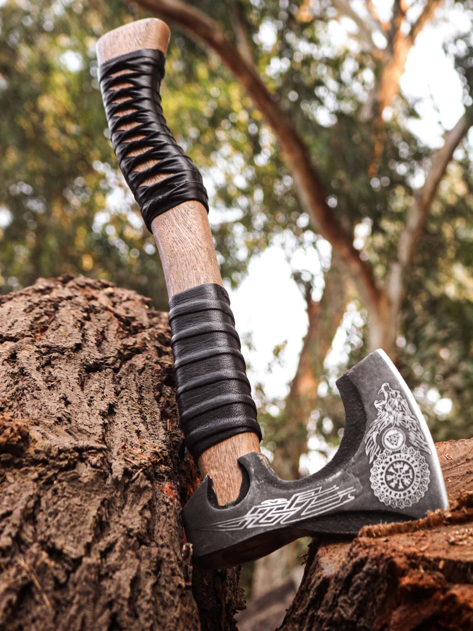 An axe with a brown wooden handle and a black leather cover over the blade, placed on a tree stump with a blurred background.