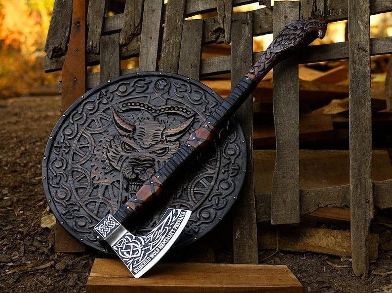 A hand-forged high-carbon steel axe with a rosewood handle, featuring detailed wolf head engraving, displayed alongside a wooden shield with a wolf design.